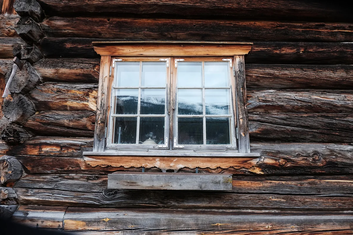 Вікно з дрібною розкладкою в стіні зі старих колод; у шибках віддзеркалюється засніжений гірський хребет